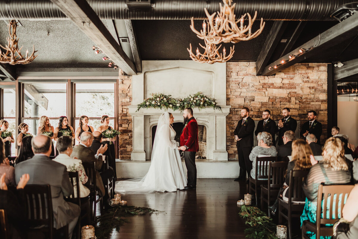 A warm and intimate wedding reception at The Lake House in Calgary, viewed from outside through large glass windows. Guests are seated at long, candlelit tables, engaged in conversation and enjoying the elegant atmosphere. The rustic charm of the venue is highlighted by stone walls, a grand fireplace adorned with greenery and candles, and antler chandeliers casting a soft glow. The dim lighting and reflections in the glass add depth to the scene, creating a cozy and romantic ambiance for the celebration.
