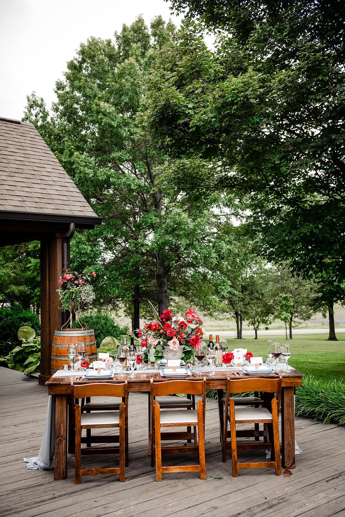 A dark wood farm table with brown wood garden chairs with ivory seat cushions set on the porch of the Stone House at Arrington Vineyards. The farm table and wine barrel are set with bright red, dark red and blush roses, protea, ranunculus, red hypericum berries, anemones and anthurium, blue thistle and dianthus. The table is set with clear crystal footed wine glasses and white square plates wrapped with teal napkins and wine cork seating cards.