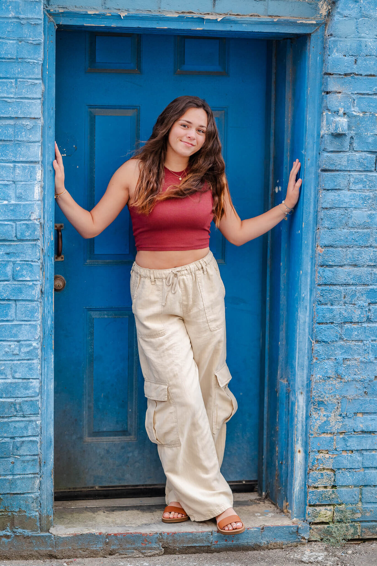 A high school senior girl with long brown hair wears khaki cargo pants and a red crop top. She is leaning in a doorway painted bright blue.