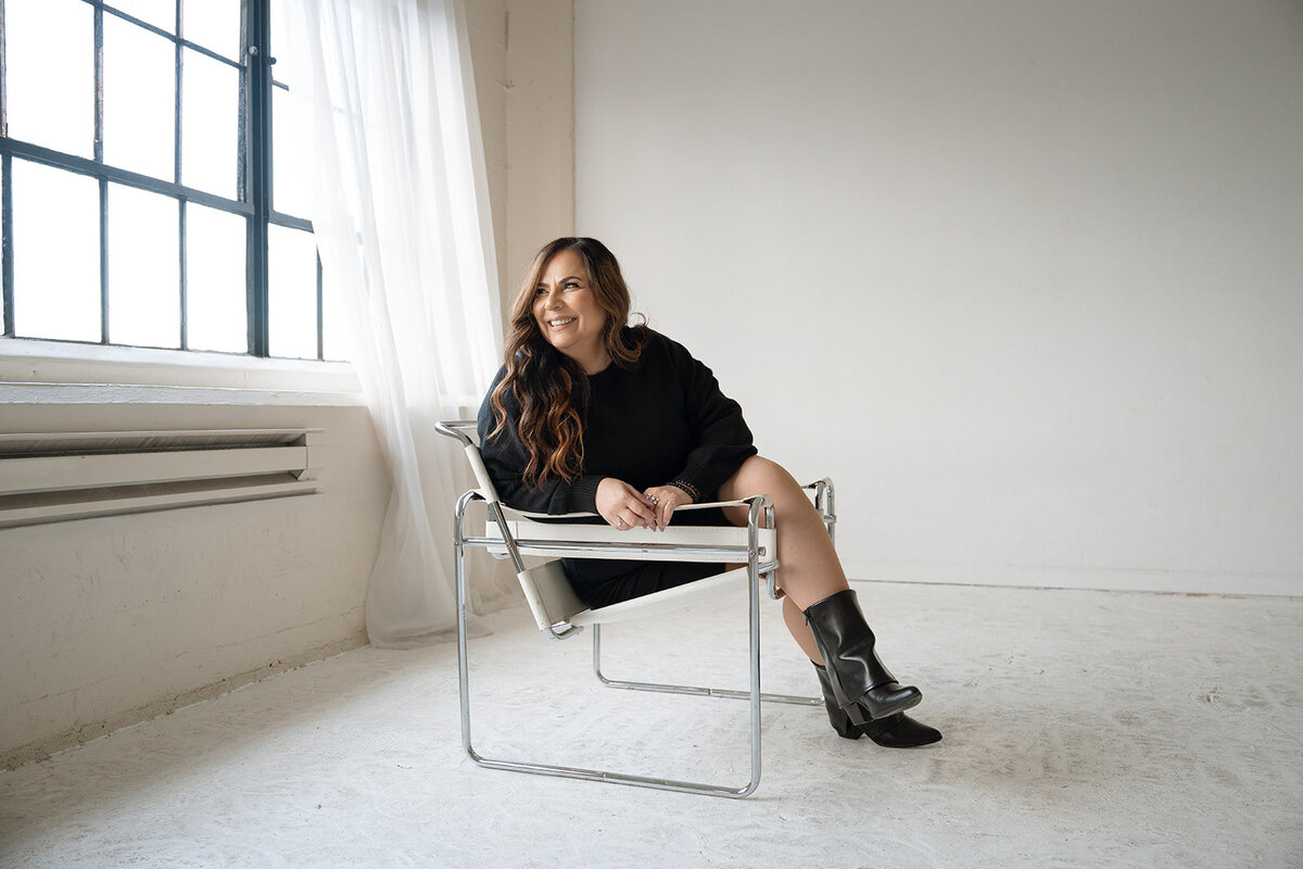 Orange County branding photo of a woman in a black outfit, sitting in a modern chair and smiling by a large studio window.