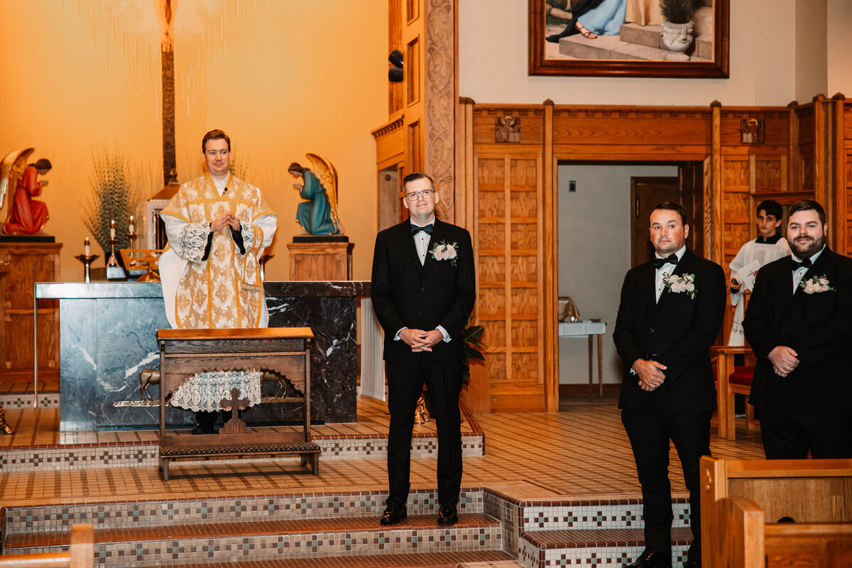 A groom in a black tux waits at the altar smiling as his bride walks down the aisle
