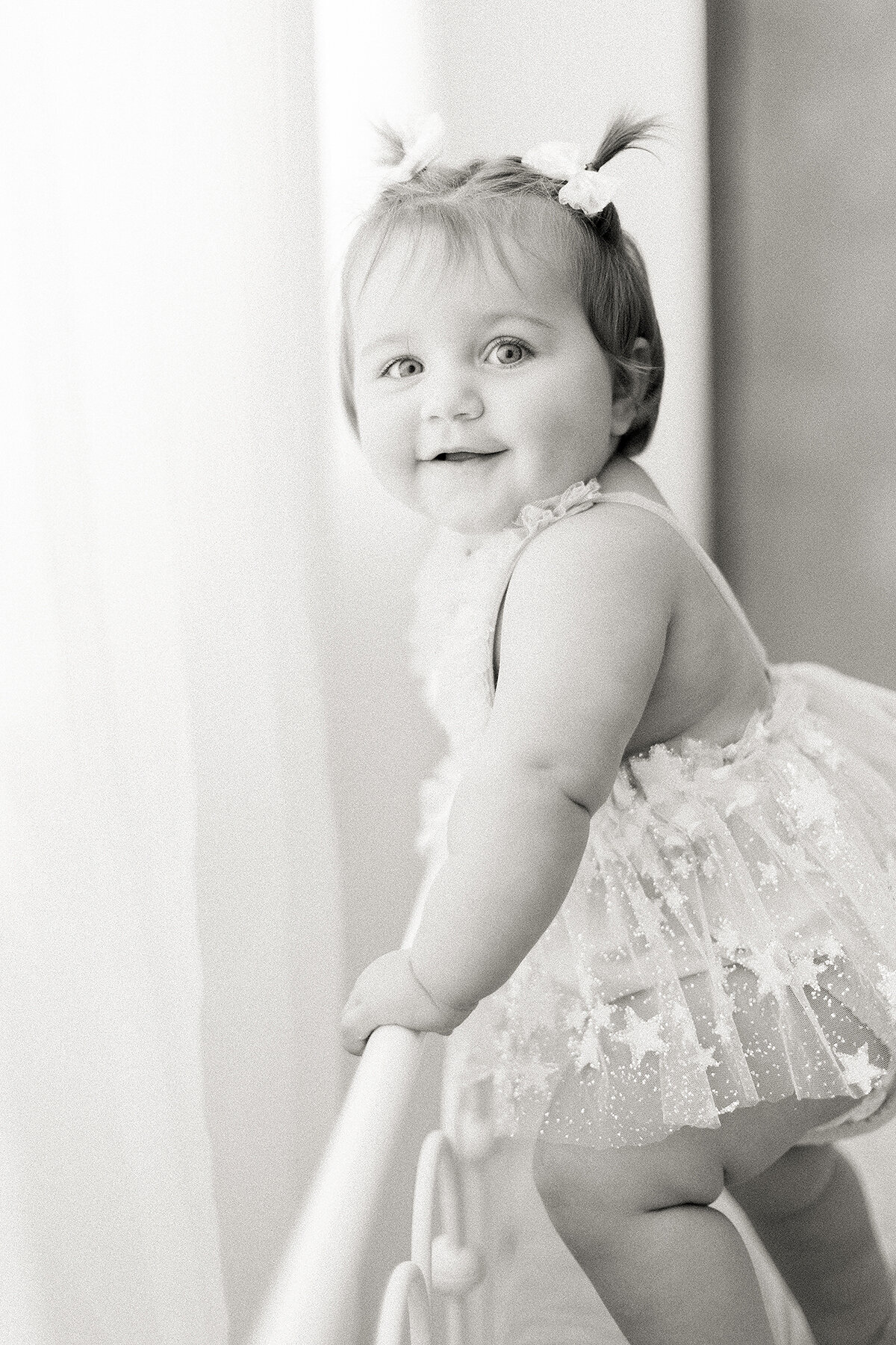 A light filled black and white portrait taken in a Fort Worth Photography Studio of a cute little baby girl standing up and holding the bed rail dressed in a tulle tutu taken by a Fort Worth Family Photographer.