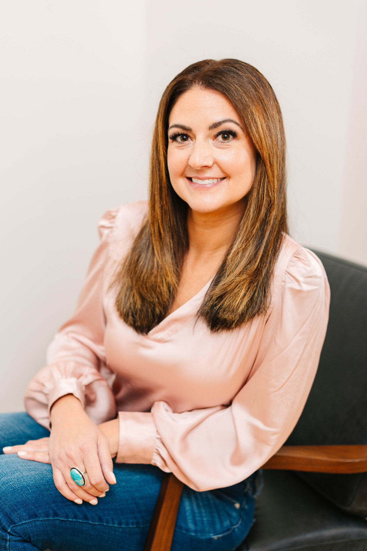 A smiling woman with long, straight brown hair sits in a black leather chair, wearing a silky pink blouse and blue jeans. She rests her hands on her lap, showcasing a turquoise statement ring. The neutral-toned background adds to the modern, professional setting
