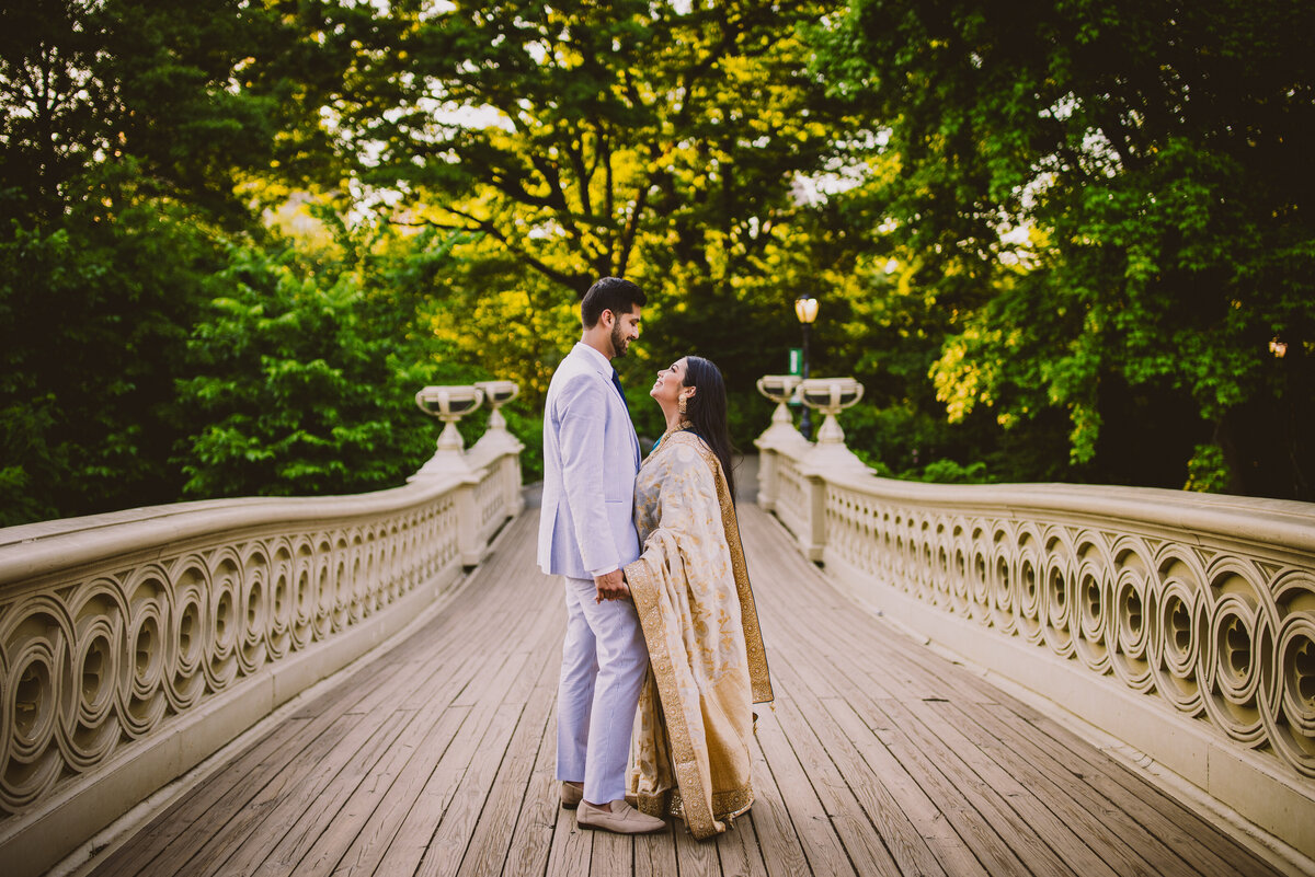 An engaged couple look at each other on the bow bridge at central park
