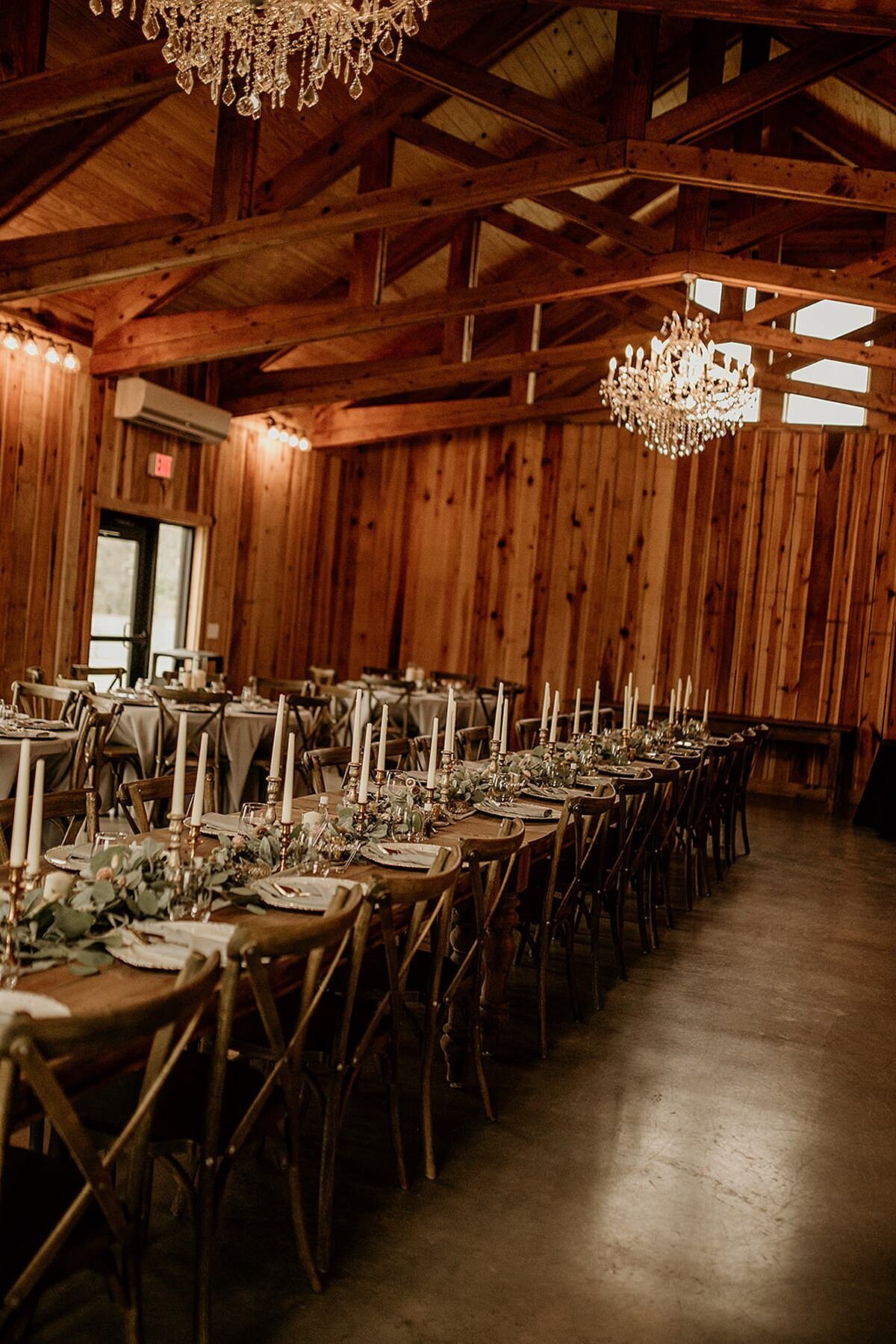 Elegant cedar wood barn with crystal chandelier has a very long row of wood farm tables with barn wood cross back chairs. The table is set with white plates a long garland of greenery and tall white taper candles in gold candle stick holders.