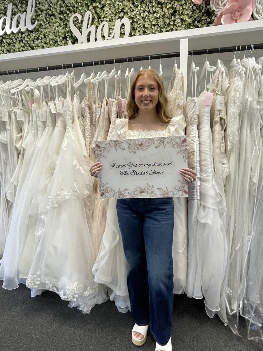 Woman standing in front of bridal gowns holding a sign announcing her dress selection