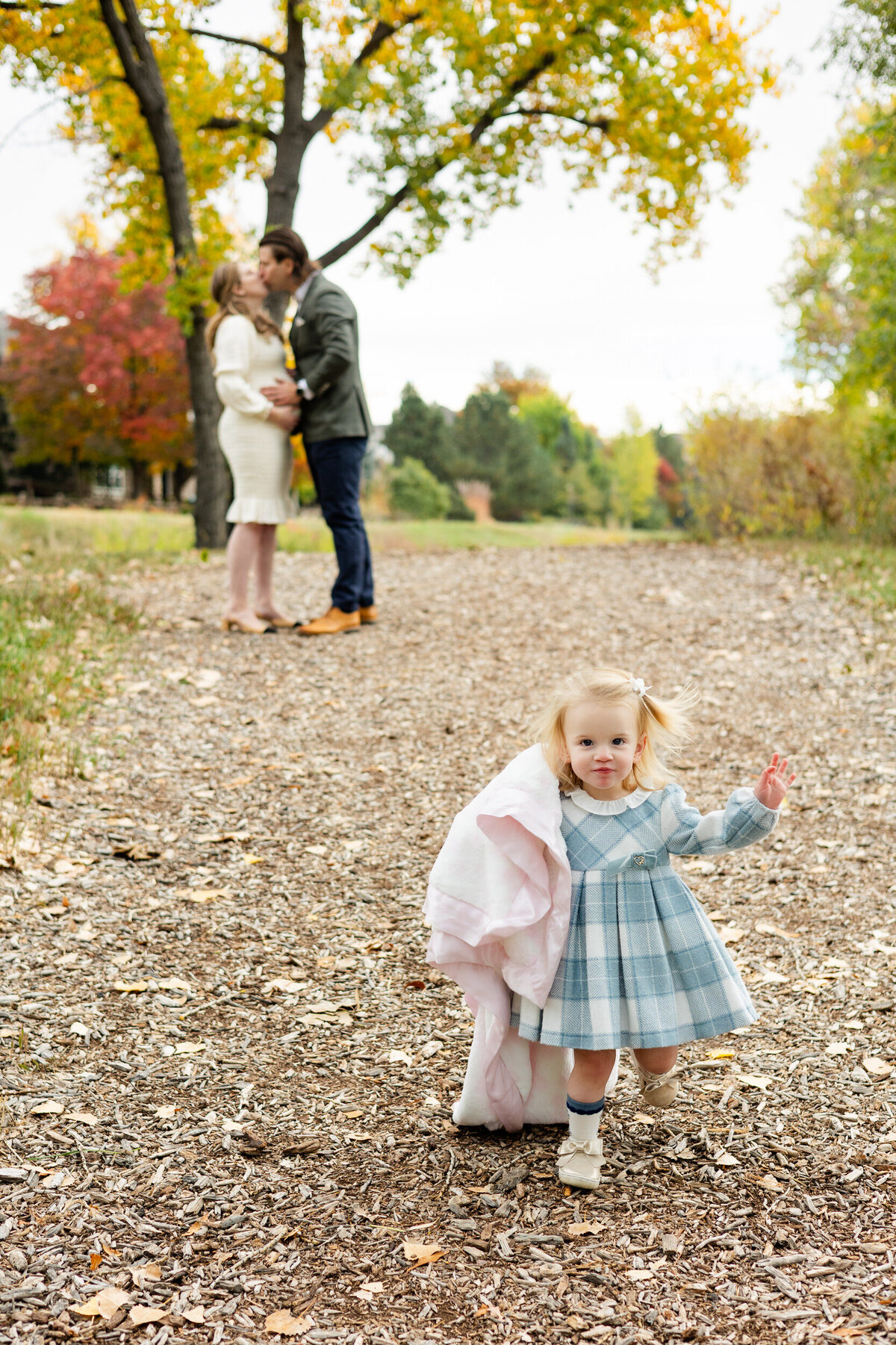 Mom and dad share a kiss in the background while toddler girl runs down the hill with her pink blanket.