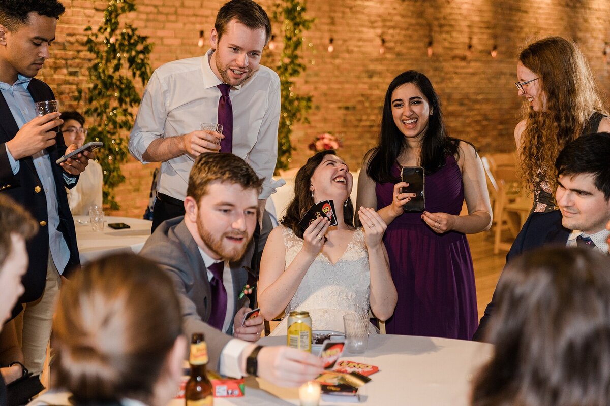 A bride and groom playing Uno with their wedding guests during their wedding reception in Plano, Texas. The bride is in the middle of the photo and is wearing a sleeveless, intricate, white dress. The groom sits to her right and is wearing a gray suit with a boutonniere. Guests sit around the table playing Uno with while other guests stand around the table chatting, laughing, and drinking.