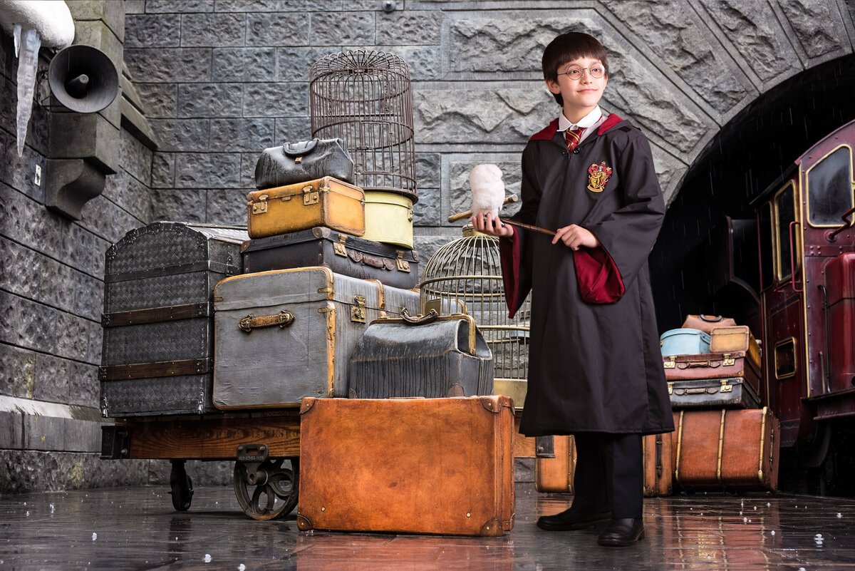 Magical fine art portrait of a young child dressed in wizard Harry Potter robes, standing next to vintage luggage in a whimsical train station setting. Captured in Overland Park, Kansas, this enchanting setup is perfect for families seeking creative and storybook-themed child photography
