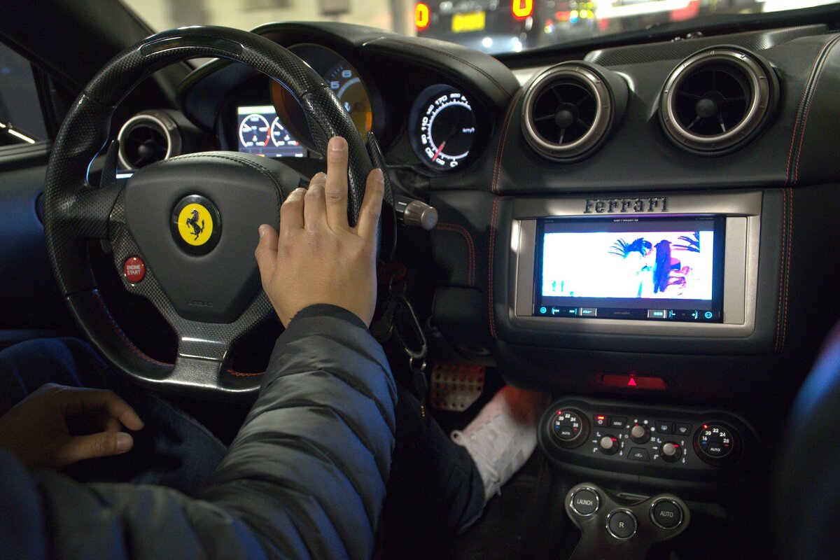 A close-up view of a person driving a Ferrari at night in Tokyo, showing the dashboard, steering wheel with the Ferrari logo, and touchscreen display, all lit up with city reflections.