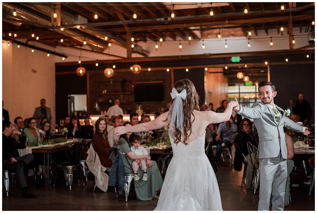 A bride with a white bow type veil has her back turned to the camera while she dances with her new husband who is faced toward the camera, at their wedding venue skylight in downtown Denver
