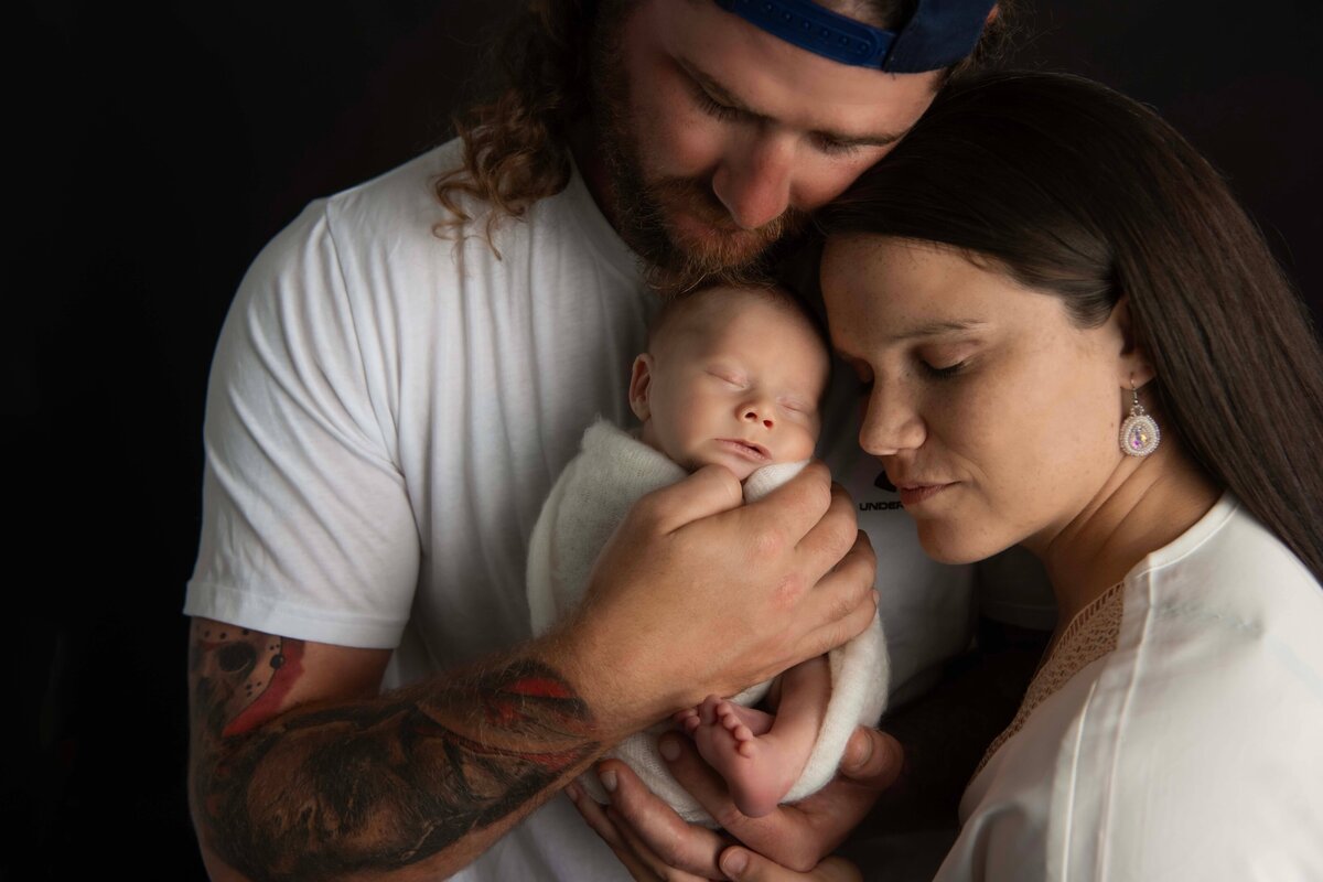 A newborn is swaddled, and is resting against his dads back for this intimate photo by Mobile, AL newborn photographer, The baby's back is against his dads chest, and he has a little foot sticking out. One of dad's hands in craddling the baby's butt, while the other is holding the baby snug against his chest. The dad's chin is resting on the baby's head, as mom comes in from the righ side and rests her head on dad's shoulder. They are all resting with their eyes closed as they soak in their new baby.