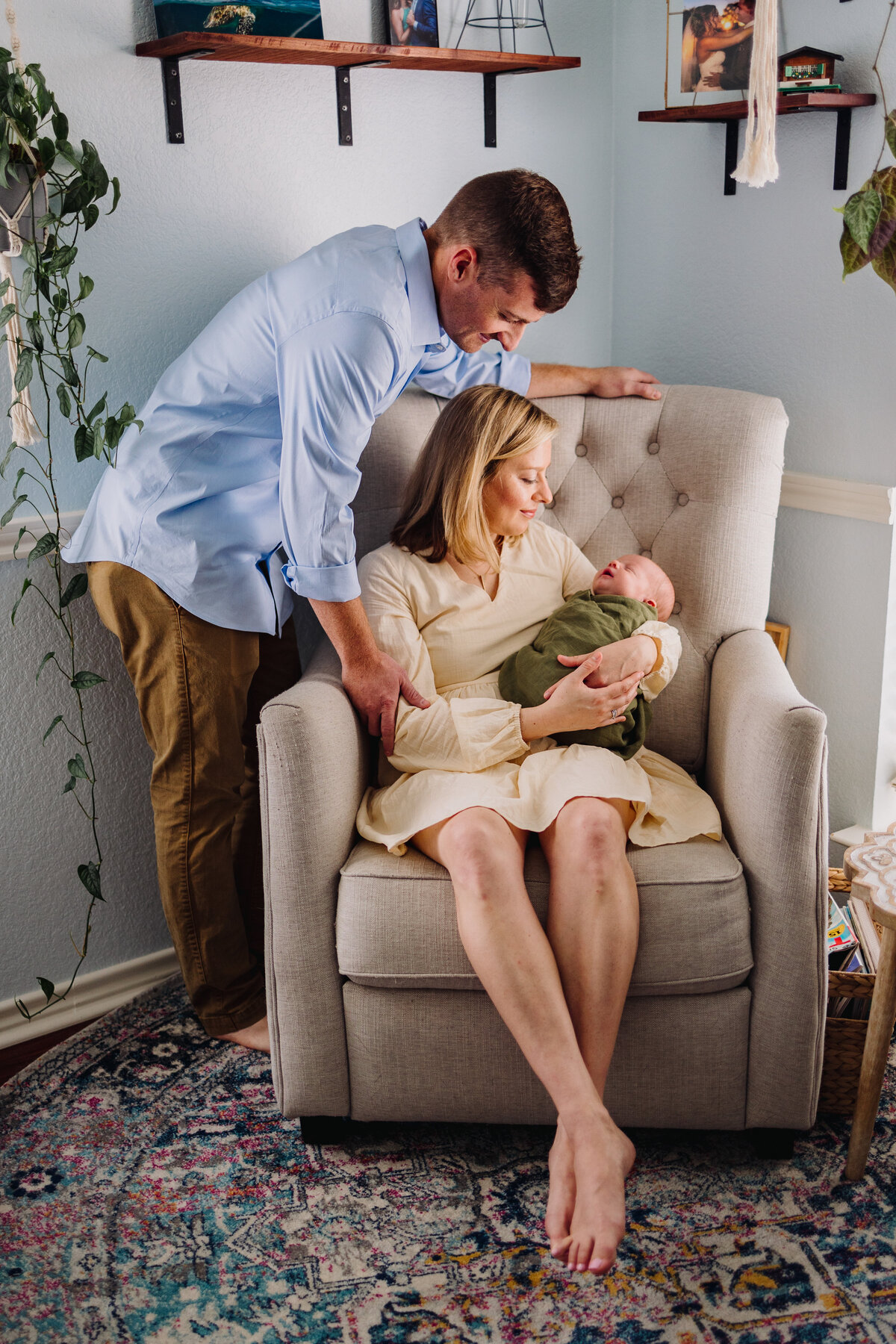 Photograph of a family with their newborn child, the woman is sitting on a white sofa and in her hands she is holding the crying baby with a green blanket. The father is looking at him and is wearing a blue shirt and brown pants