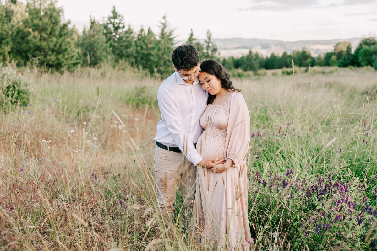 Beautiful portrait of a woman and her husband in an open field with their hands on her pregnant belly. They are cuddling and looking down at her belly.