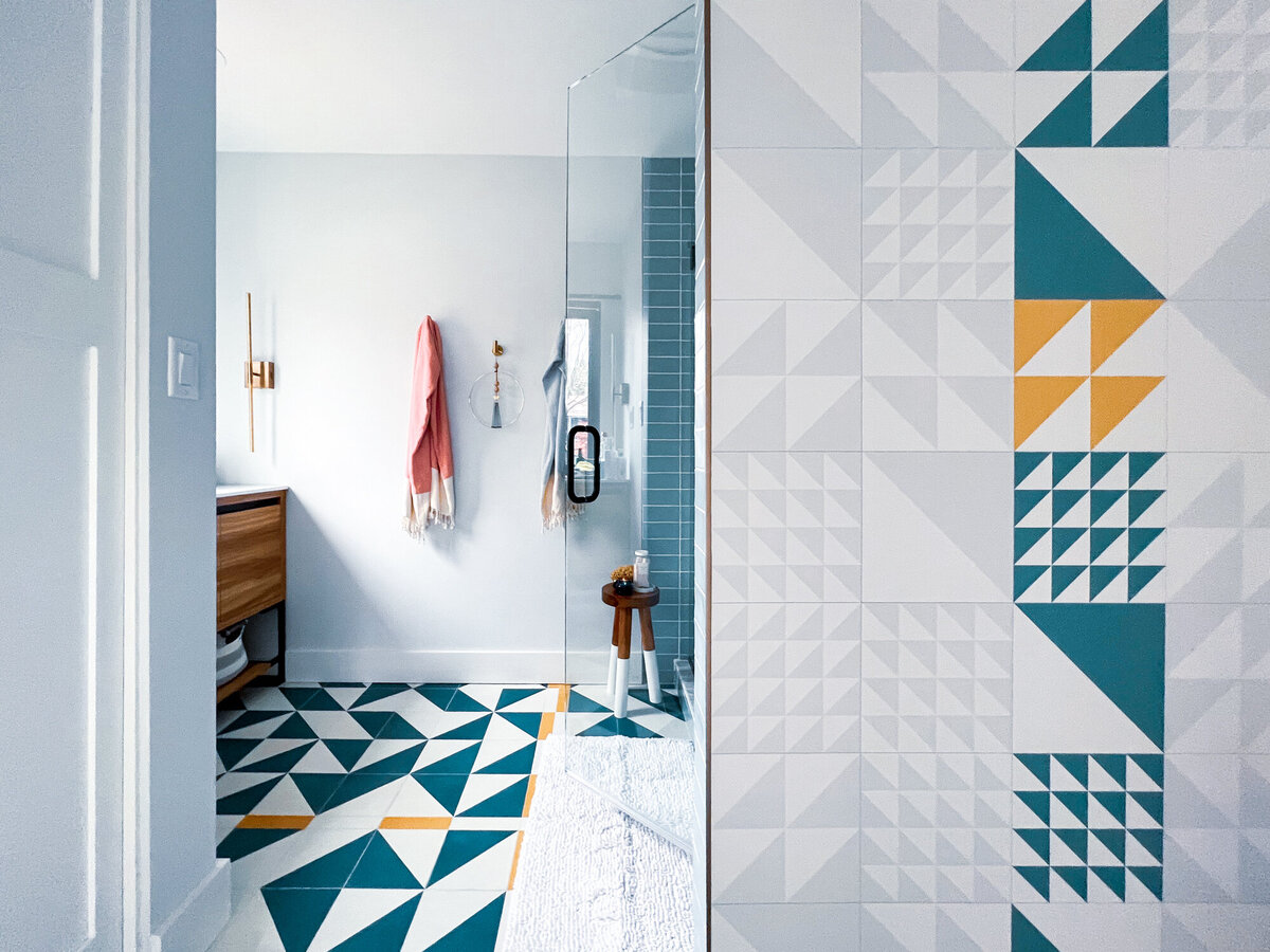 A brightly lit, modern bathroom with geometric patterned tiles in teal, white, and yellow covering the floor and extending up a portion of the wall. A clear glass shower enclosure is partially visible on the right, with a small wooden stool inside. A light blue tiled wall is visible inside the shower. Outside the shower, a white wall features a wooden vanity with gold hardware, a gold towel bar, and a coral-colored towel hanging from a hook. Another towel hangs from a hook near the shower entrance. The bathroom door is partially visible on the left. The geometric tile pattern on the floor appears to be the "Prism" pattern, creating a dynamic and contemporary look.