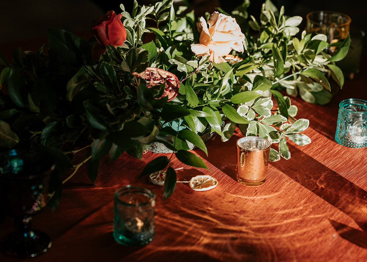 A rich floral centerpiece with roses, greenery, and dried citrus slices on a wooden table surrounded by teal and gold candle holders