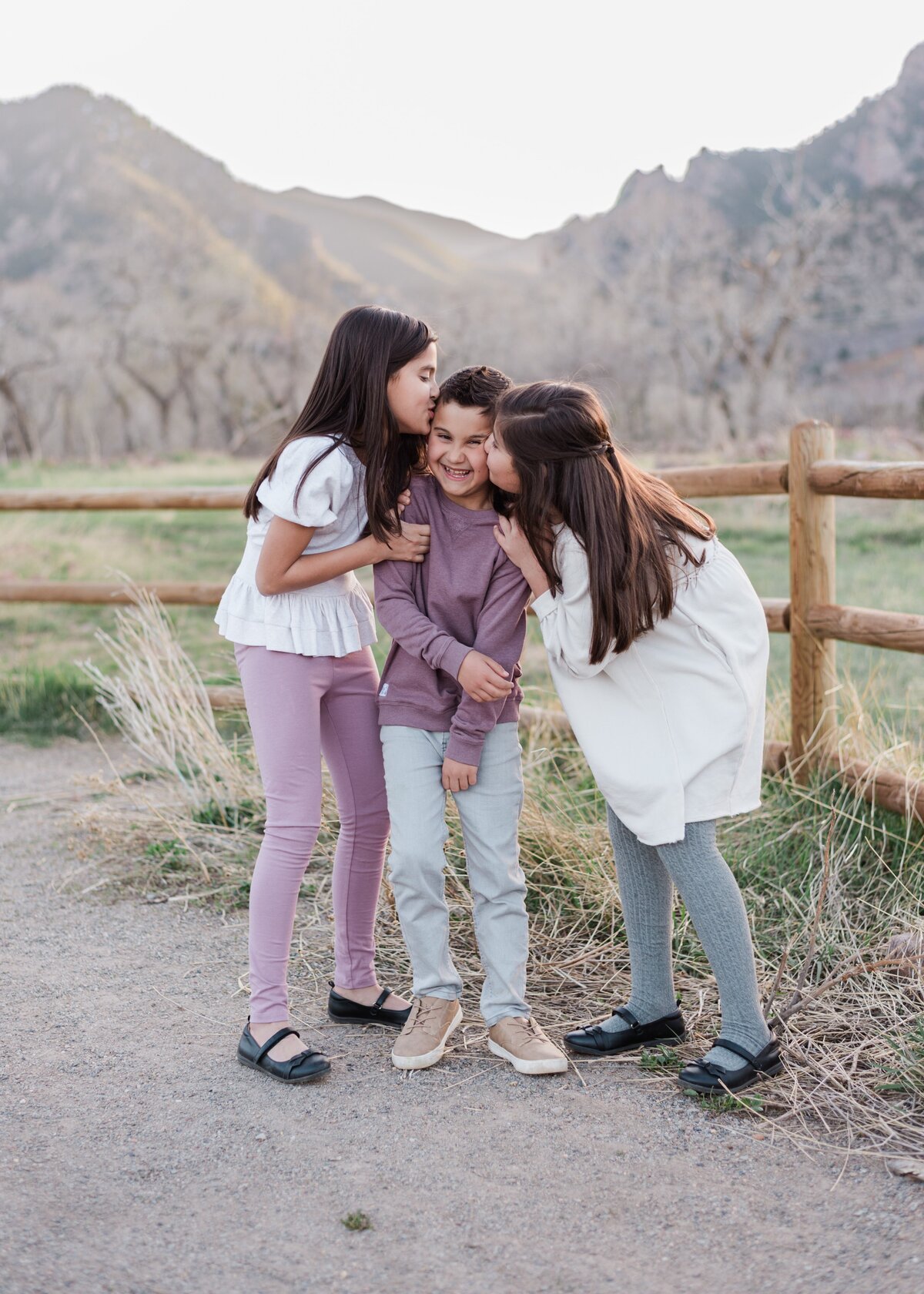 Two young girls in white tops kiss their brother in a purple sweater on the cheeks as he smiles and squirms with the boulder flat irons in the background