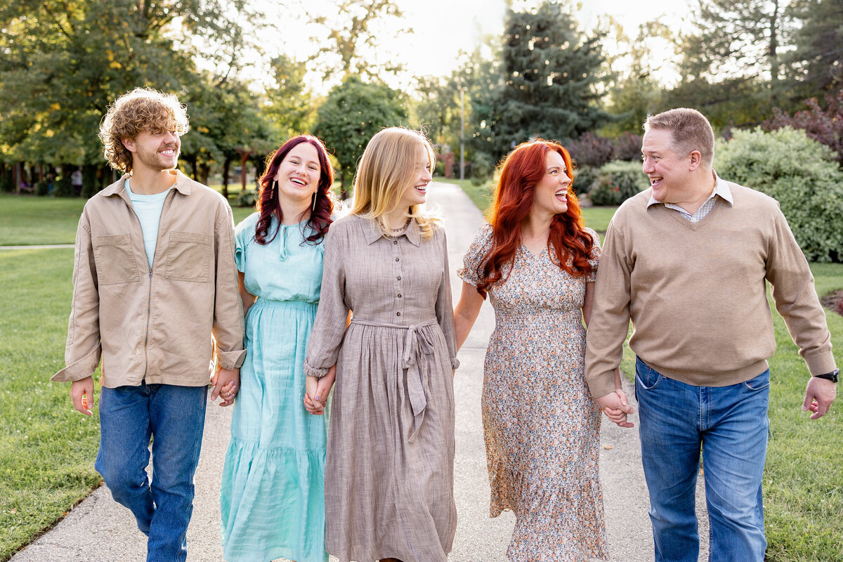 A family of five walks hand in hand down a scenic pathway, sharing smiles and laughter in a lush green park.