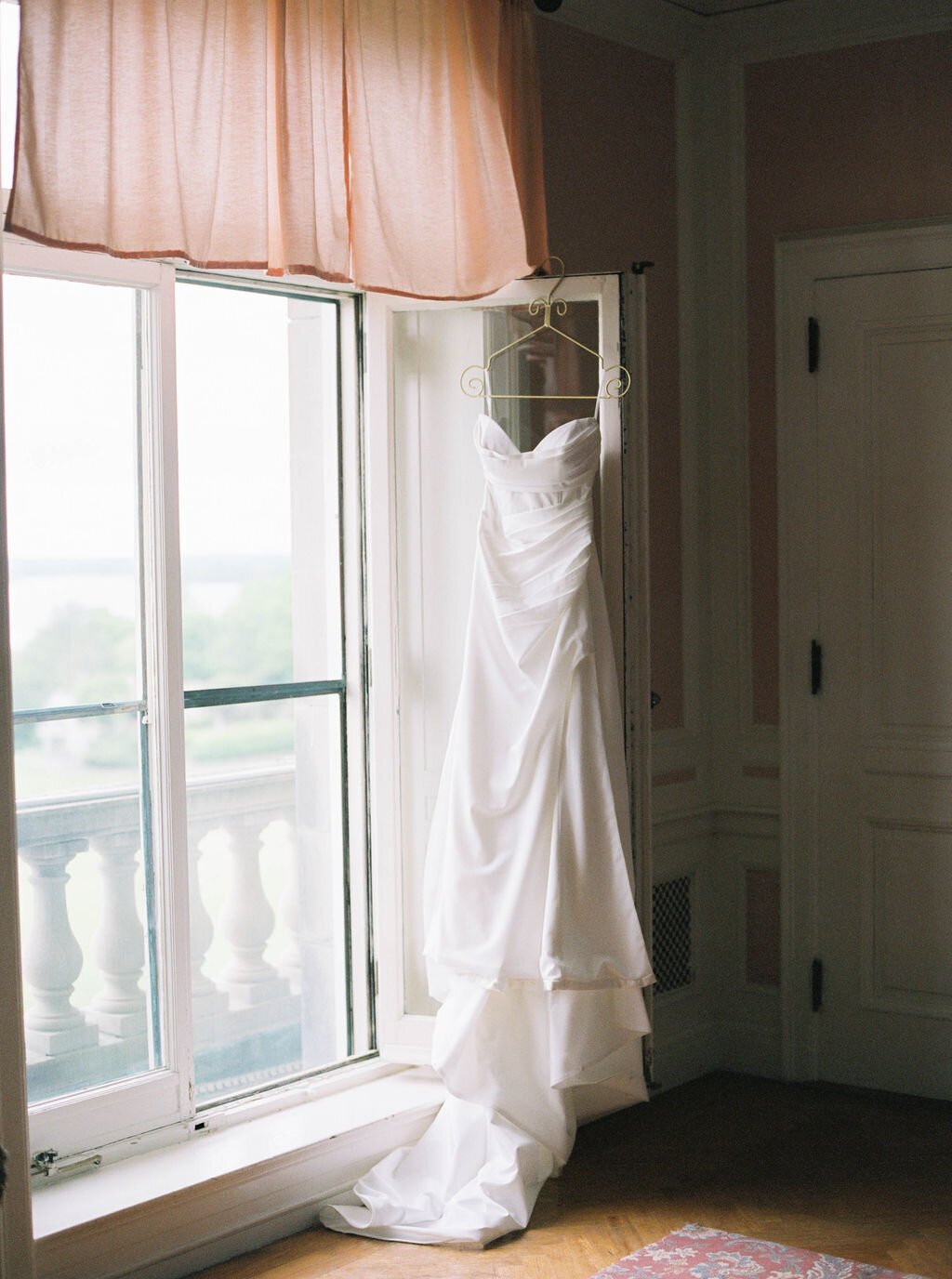 Newport Wedding Photographer | White wedding dress hangs on a hanger by a bright window with pink curtains, overlooking a serene balcony.