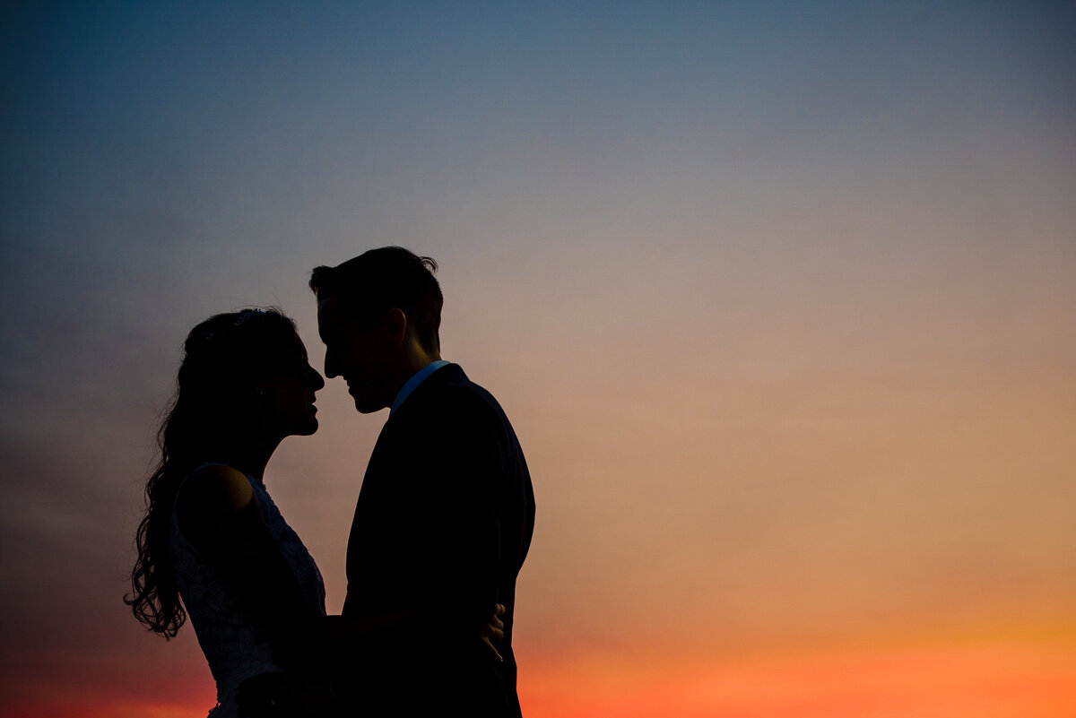 Silhouette of a couple with their faces close to each other in front of a blue and orange sunset by Charlotte wedding photographers DeLong Photography