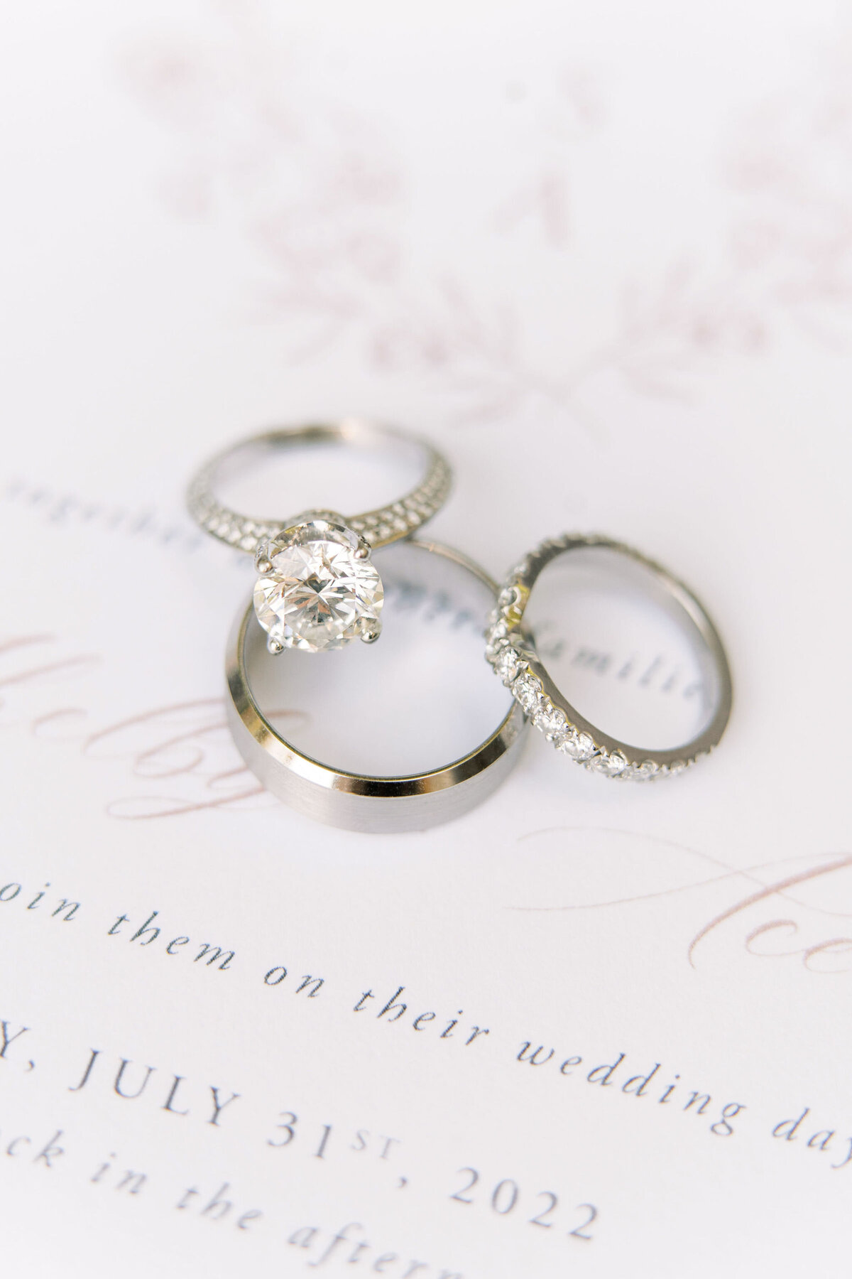 Three rings, including a diamond engagement ring, a wedding band with small diamonds, and a plain wedding band, rest on a wedding invitation card, perfectly showcased by our expert Banff wedding planner.