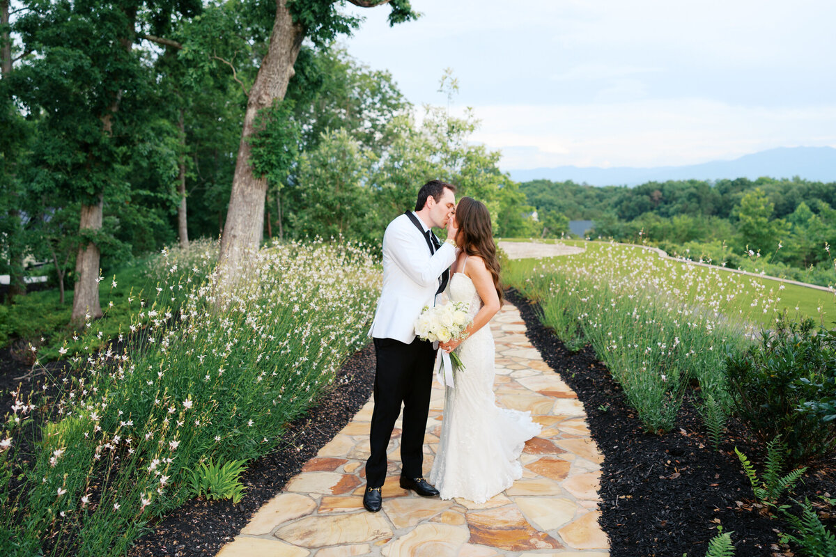 Bride and groom sharing a kiss on a stone path surrounded by wildflowers, with mountain views in the background at their outdoor wedding in Tennessee