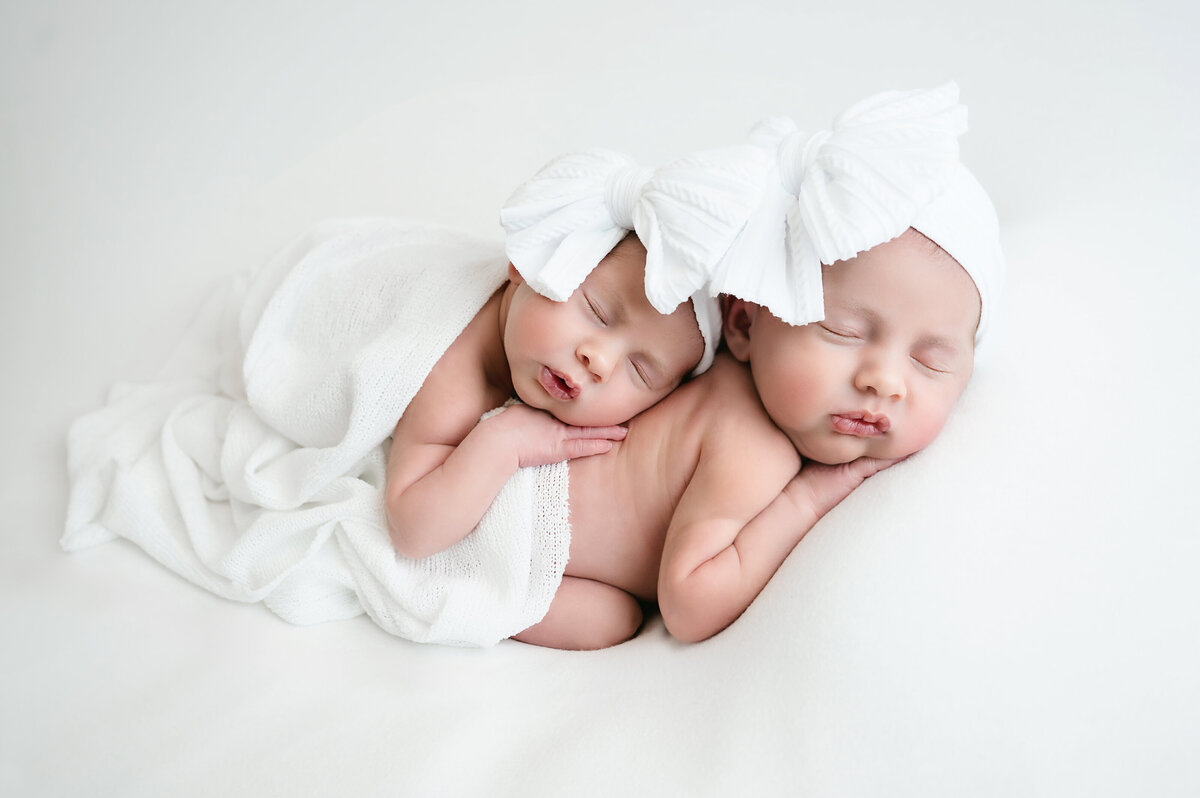 A precious twin newborn photography session in Pittsburgh, capturing two sweet babies bundled in soft white blankets with matching large white bows. This serene image celebrates the beauty and tranquility of newborn twins during their first days
