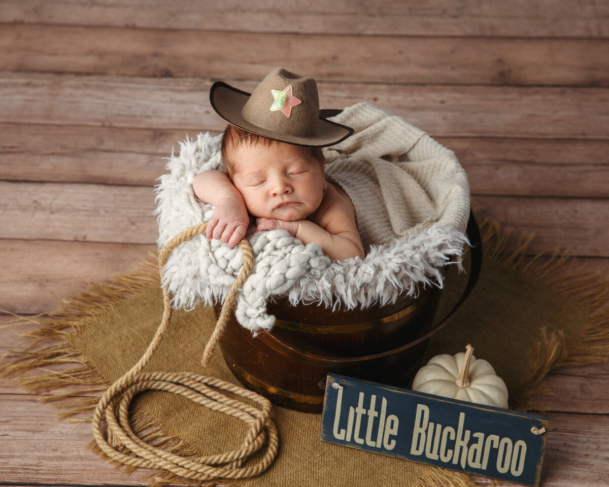 Cute portrait of a tiny newborn baby posed in a bucket with a tiny little cowboy hat and a rope