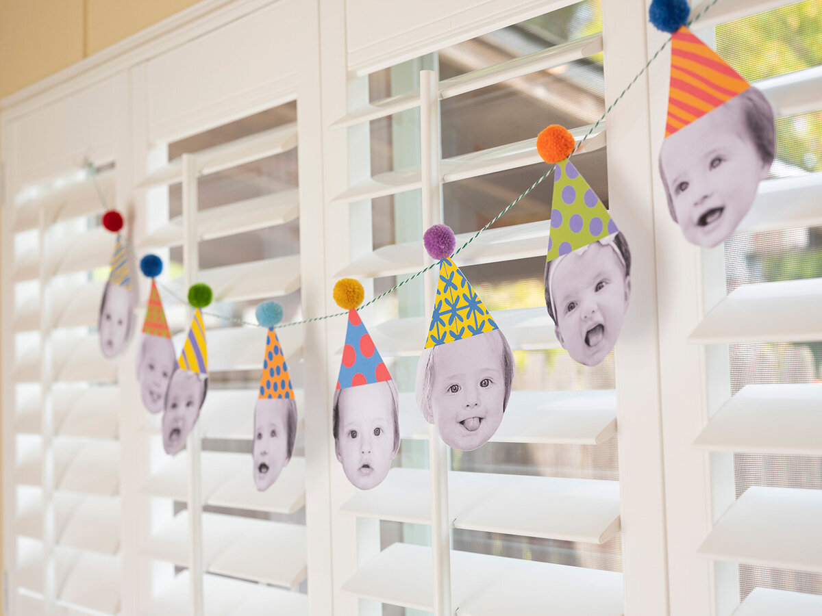 Garland of baby’s faces wearing birthday hats hanging on a window as backyard party decor for a first birthday celebration in the Bay Area.