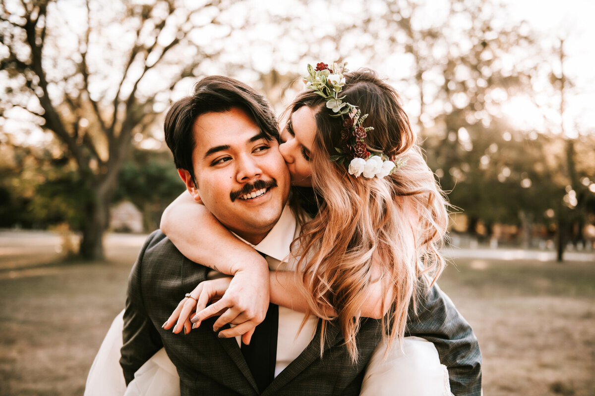 Couples Photography, Woman with light pink hair is on the back of a man with a mustache as she kisses the side of his face.