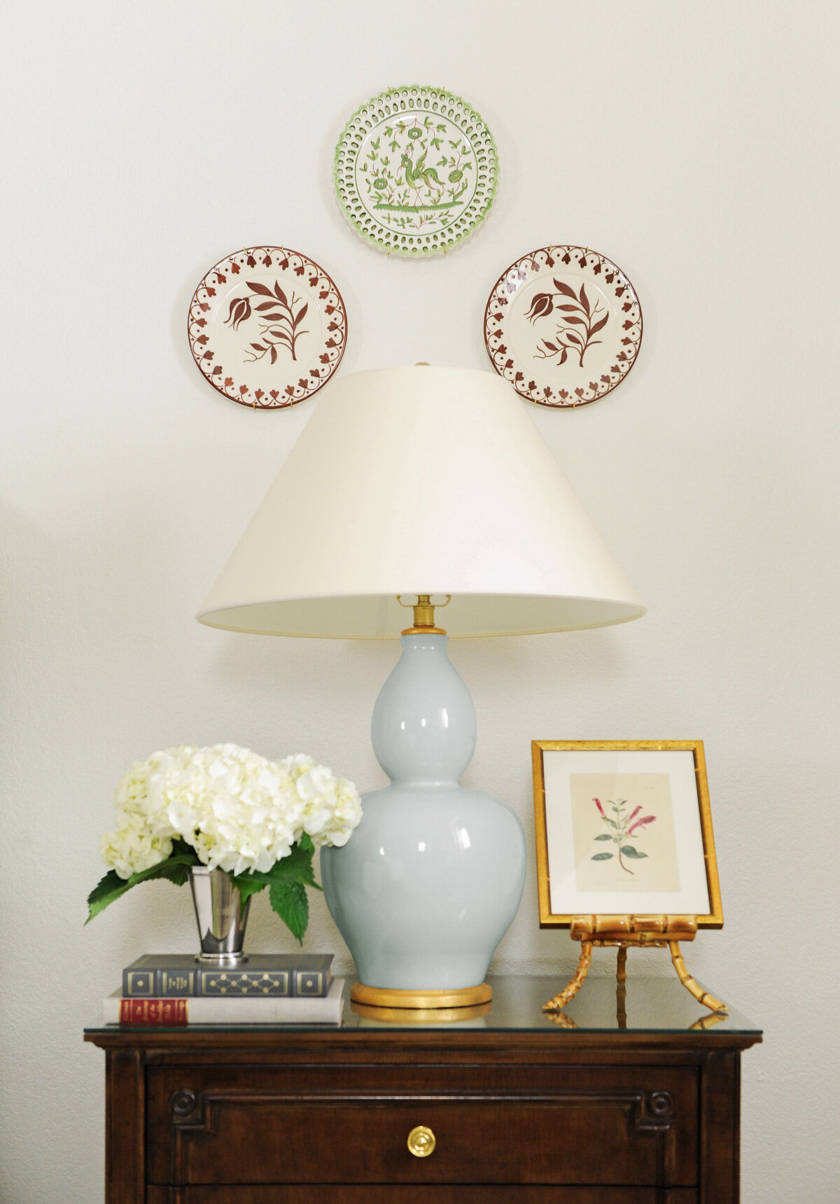 Blue visual comfort nightstand lamp on wooden nightstand with white hydrangea floral arrangement and bamboo art stand. Decorative plates are used  as artwork and wall decor.