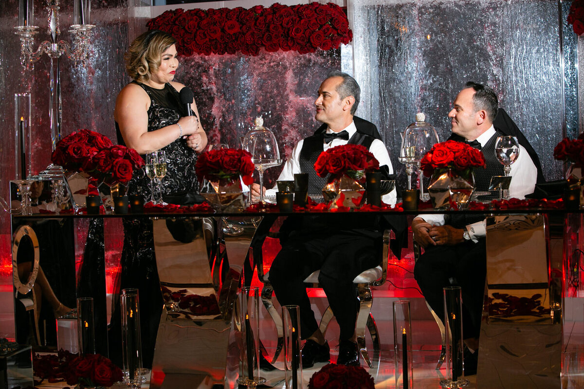 A person standing next to grooms sitting at a table and giving a speech