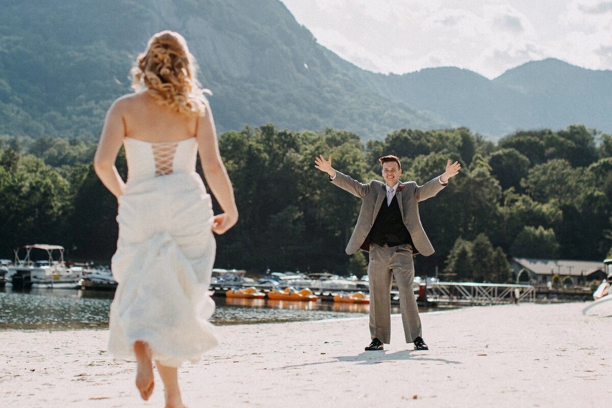 A bride runs towards a groom on a sandy beach, with the groom standing with open arms