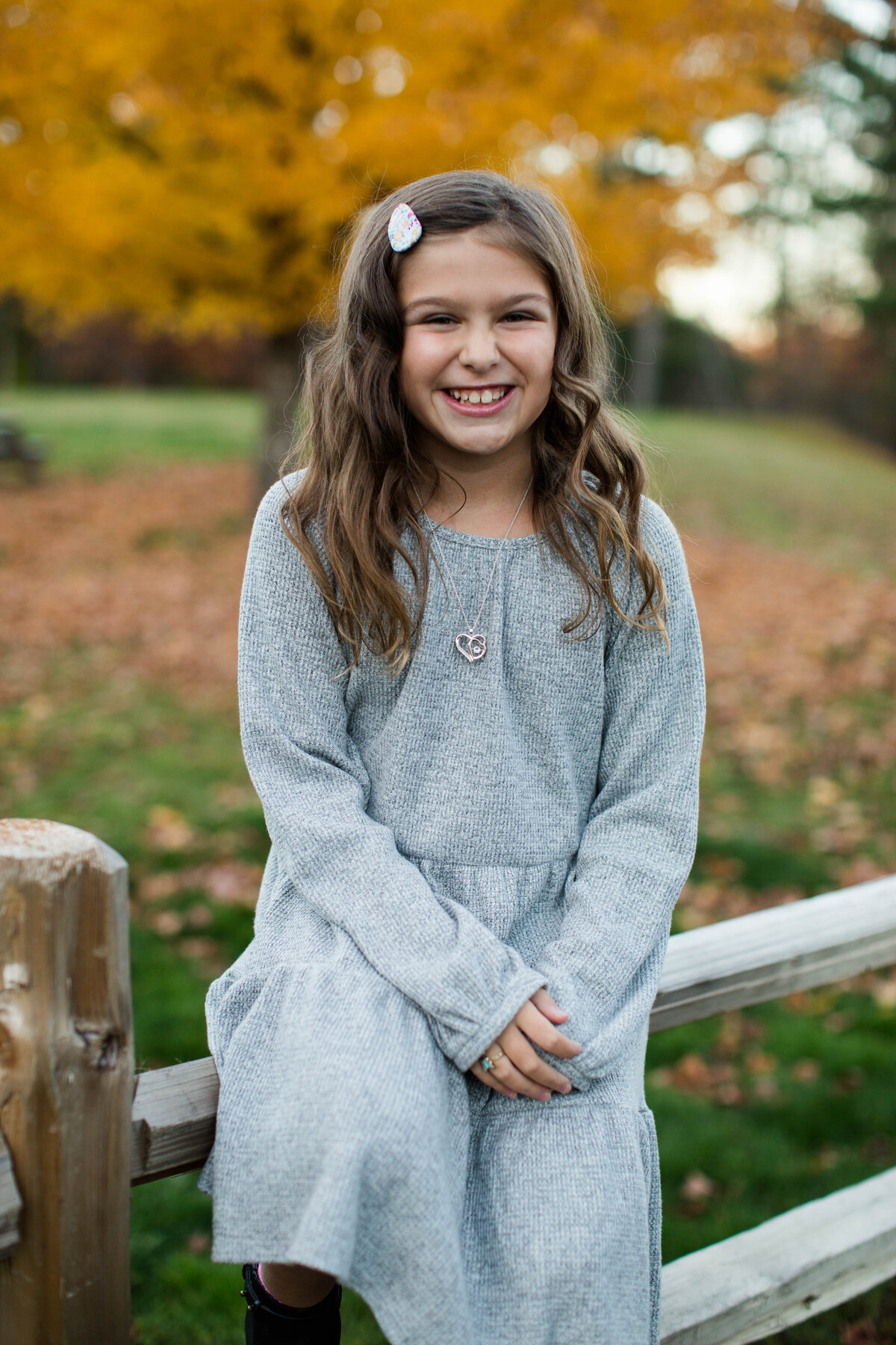Young girl in a gray dress sitting on a rustic fence smiling in the fall