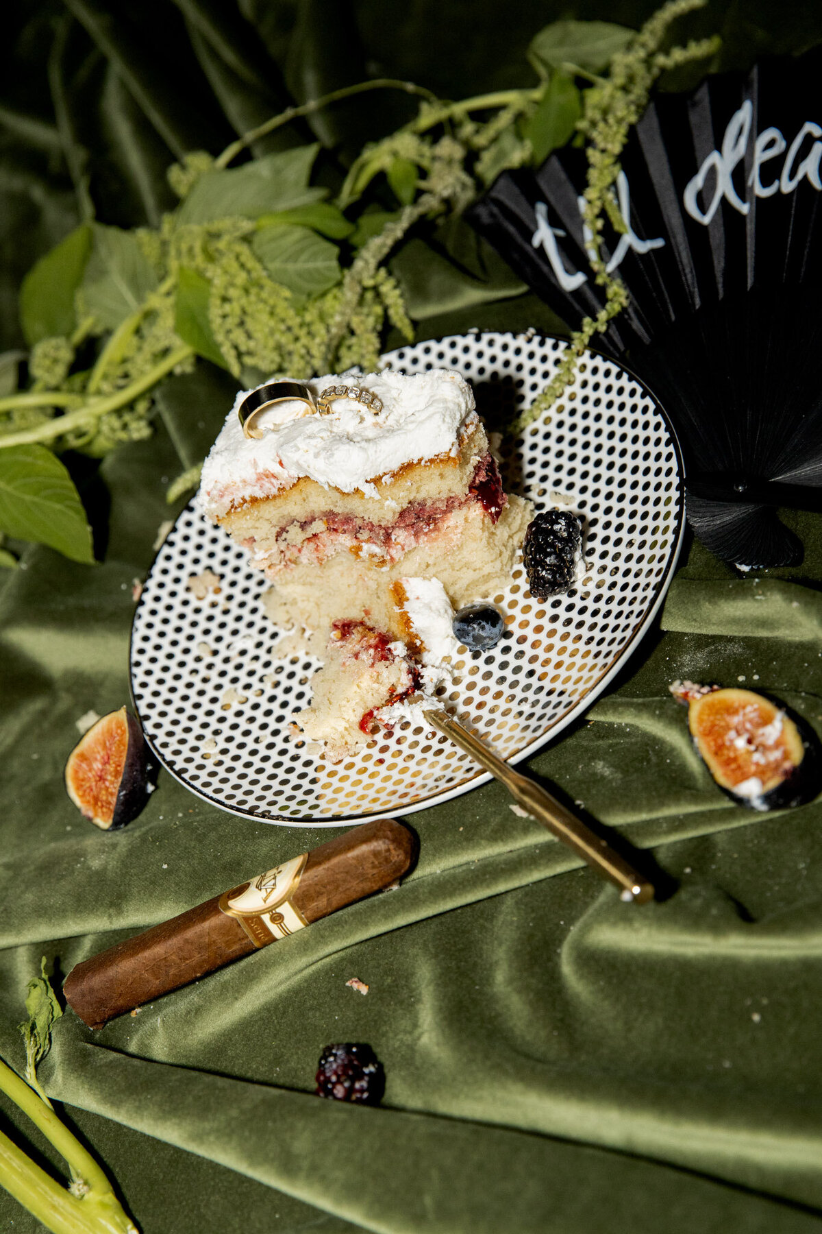 The couple's wedding rings placed in a piece of their wedding cake, with some of their wedding details, like a cigar and a hand fan, and some fruit on the plate. Editorial direct flash vibe.