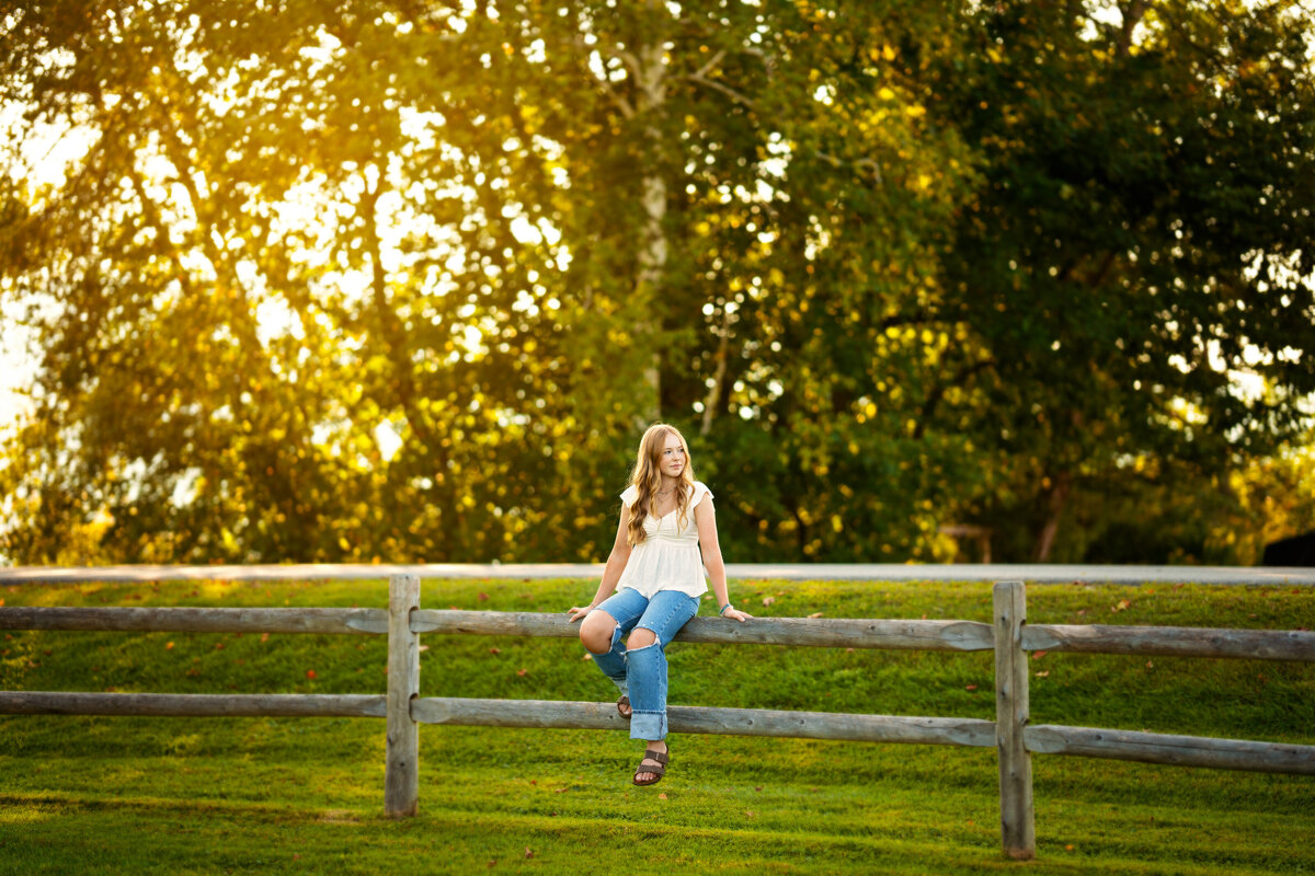 High school senior girl sitting on a wooden fence at sunset.