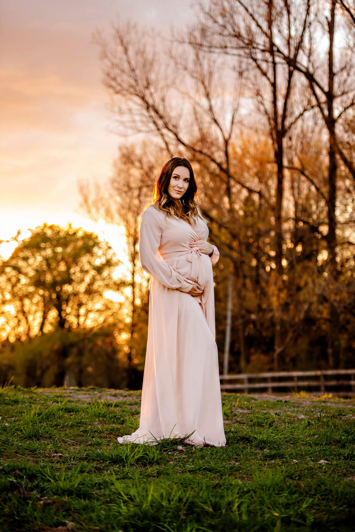 pregnant woman wearing a pink dress with the golden sun setting behind her for her outdoor summer maternity photography session in Burlington