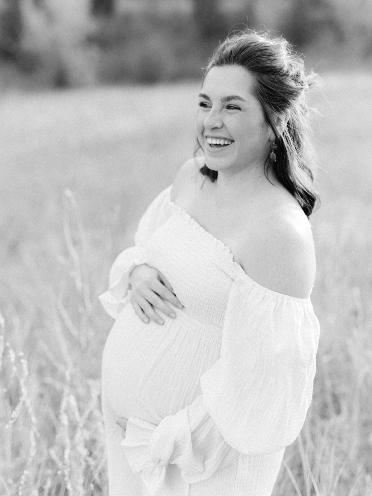Black and white photo of a woman smiling in a file d holding her baby bump- photo by Maegan R Photography