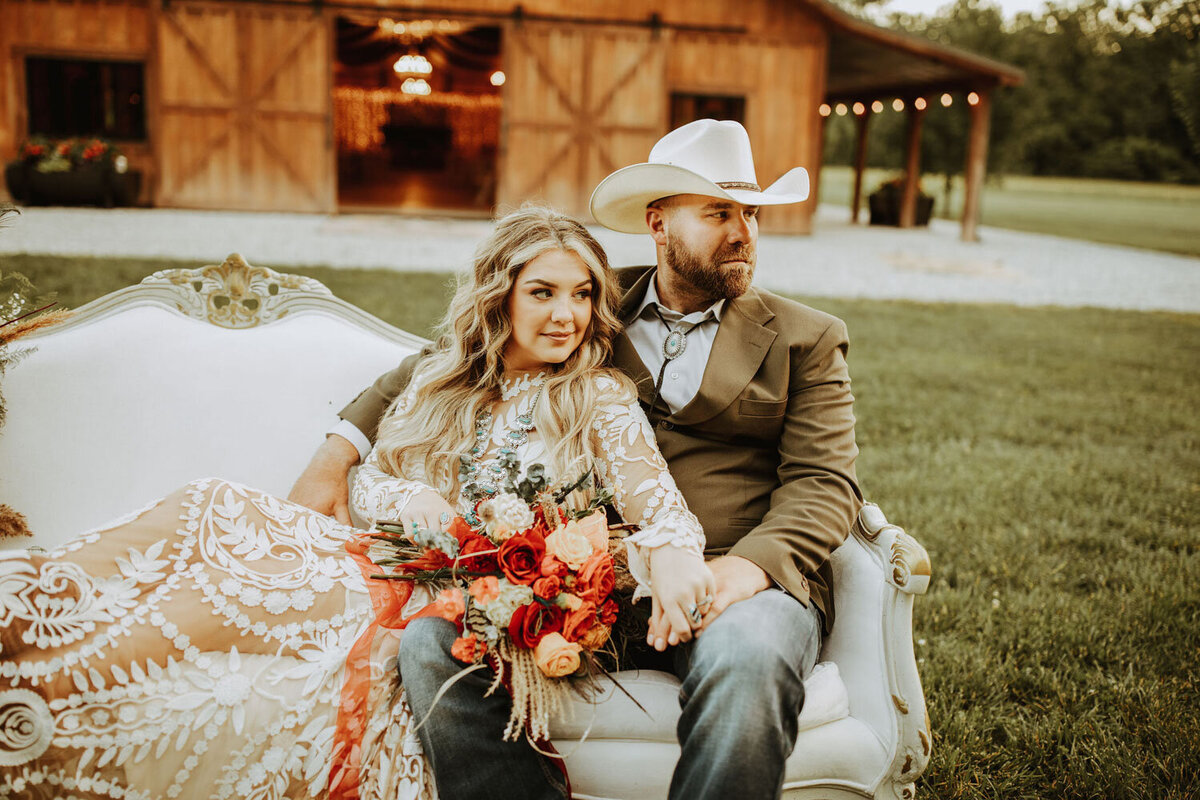 Couple sitting on a vintage couch, posing with a bouquet in front of barn