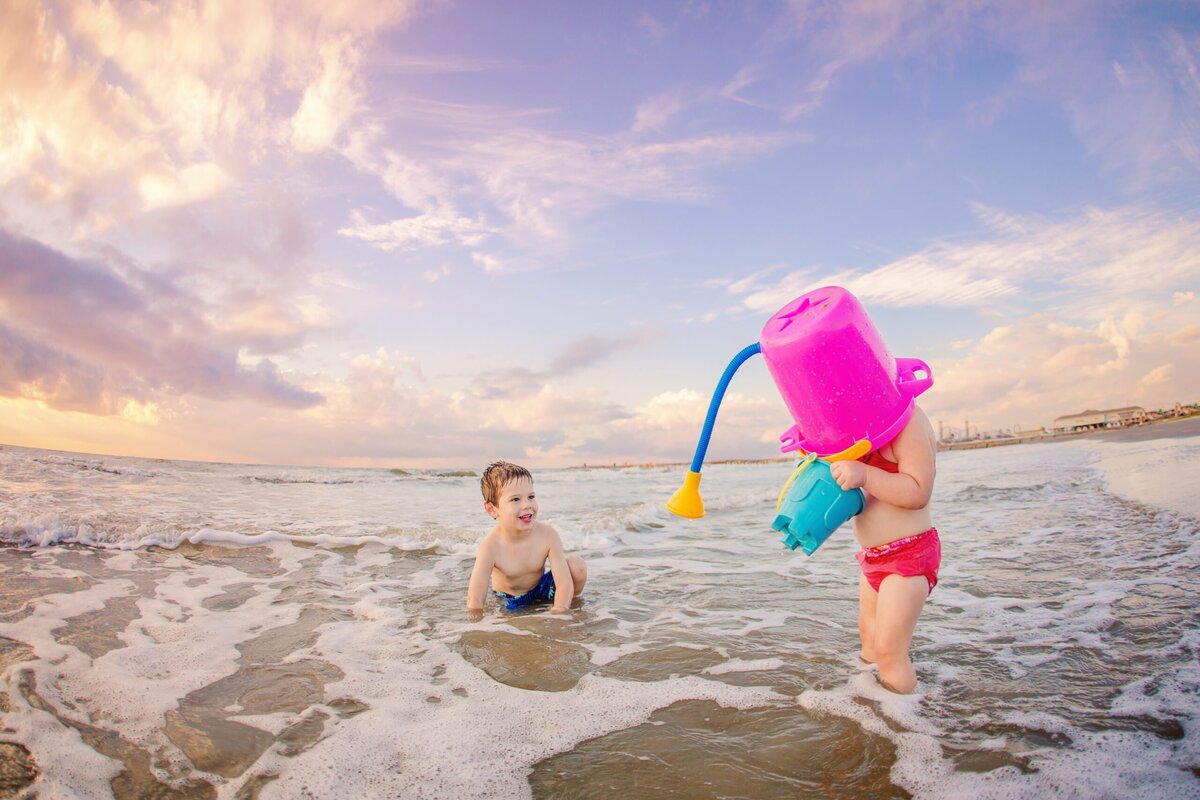 Two children play in the ocean at the beach. One has a bucket over her head.