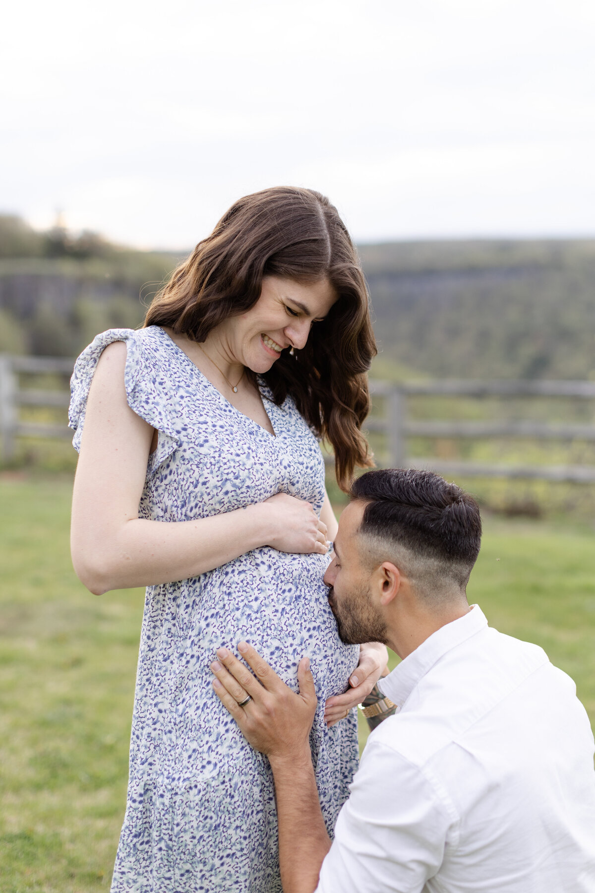 Soon to be parents are in a field as the dad is on one knee kissing the baby belly