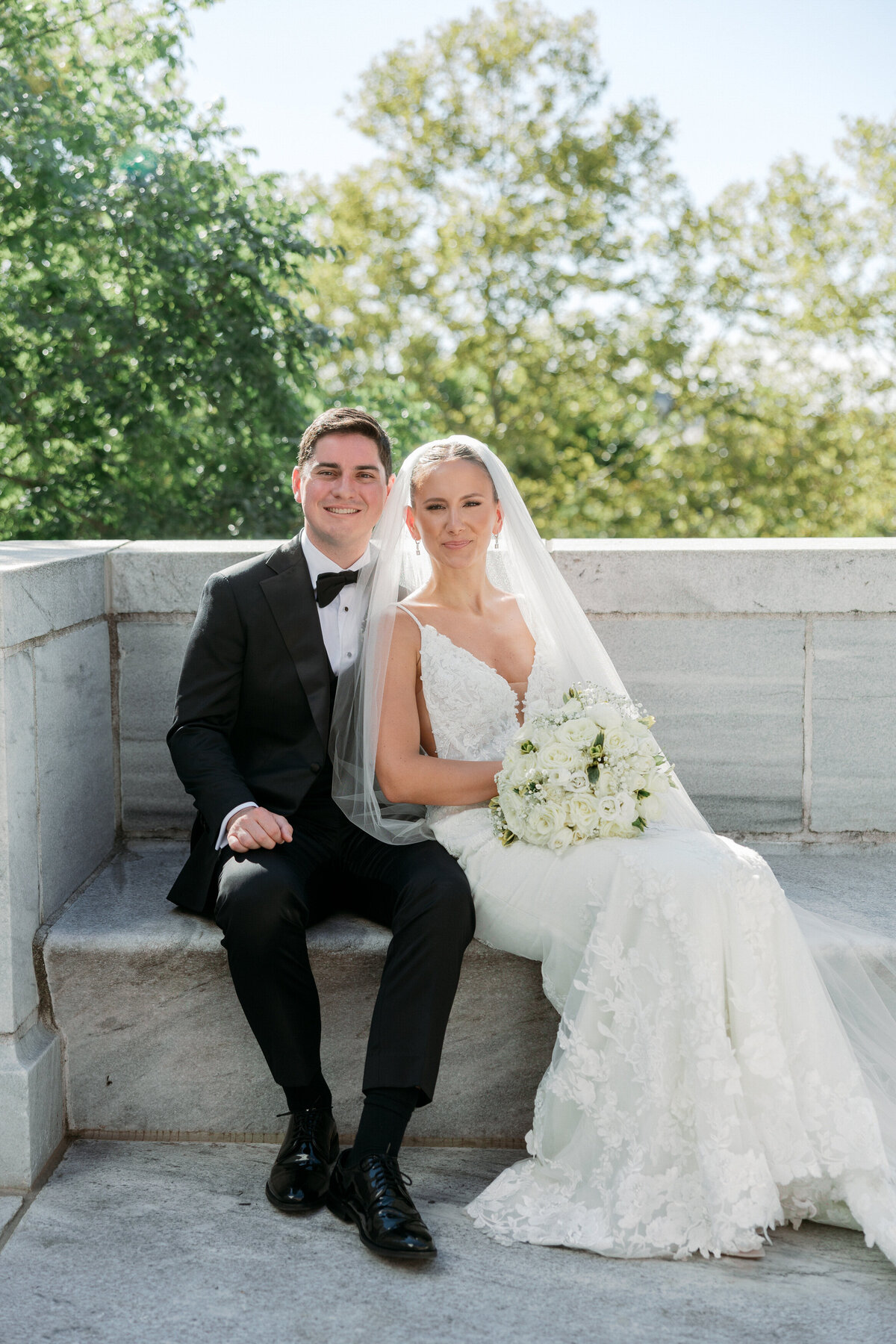 Newlyweds sitting together, smiling and holding a white bouquet with green trees in the background in Philadelphia.