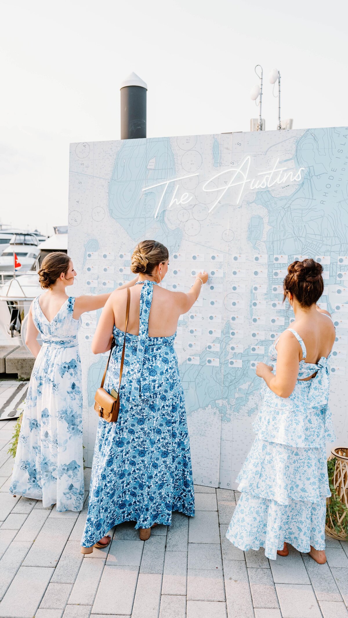 Wedding Photographer RI | Three women in floral dresses examine a seating chart at an outdoor event by the water. Boats and "The Austins" sign are visible in the background.