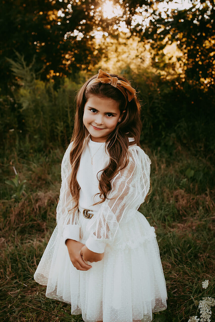 a six year old girl holding her thumbs in a white dress in a field at sunset