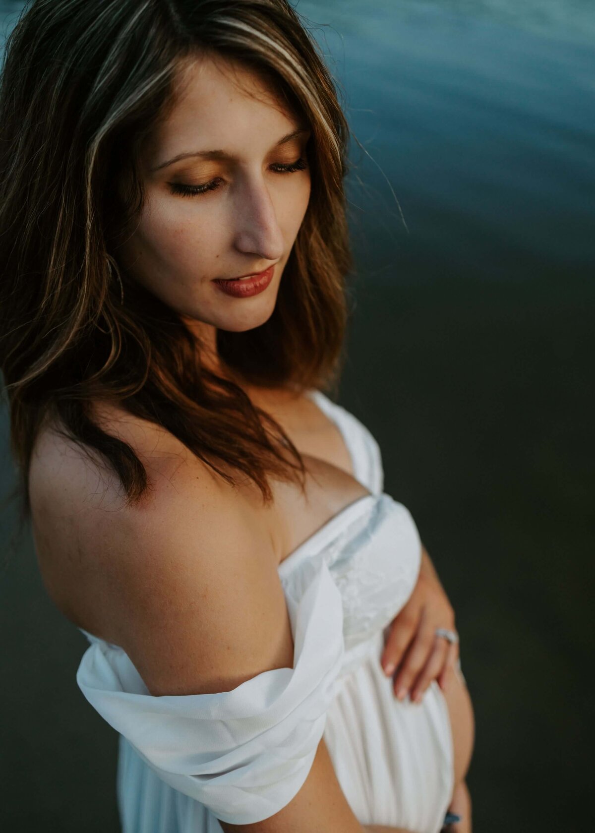 A pregnant woman in a white dress stands gracefully by the water. Embodied with the essence of Pittsburgh maternity photography.