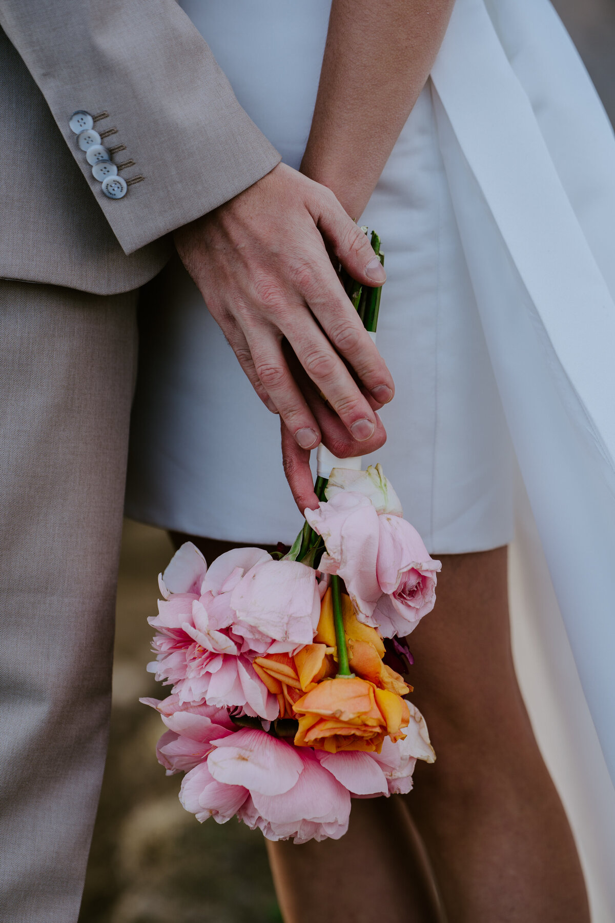 Close-up of bride holding pink and orange bouquet with groom’s hand