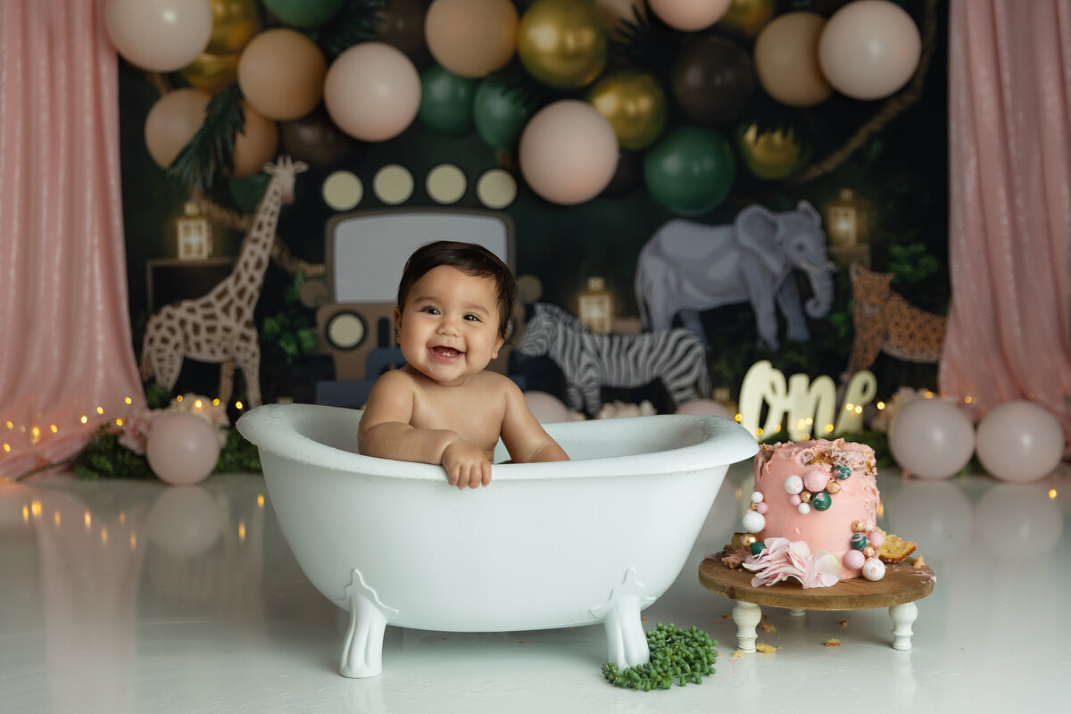 A happy toddler girl sits in a bathtub after smashing her cake for her first birthday
