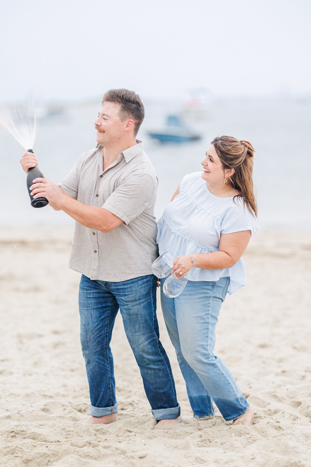 Man popping a champagne bottle on the beach while the woman smiles beside him holding stemless glasses, with blurred boats in the background at Chatham Bars Inn in Chatham, Massachusetts — representing the portfolio of a Cape Cod engagement photographer.