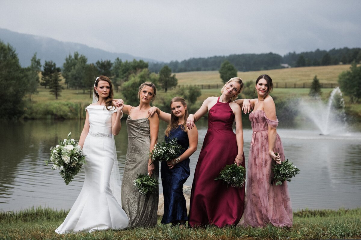 Bride and bridesmaids in mismatching dresses pose together with all white and greenery bouquets at Spruce Mountain Ranch in Colorado.