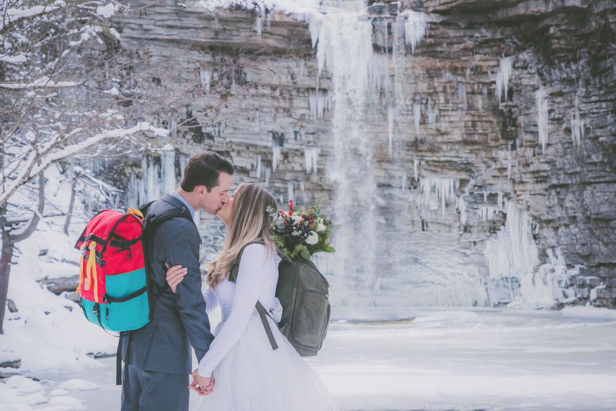 Couple holds hands and kiss with waterfall in background while wearing backpacks.